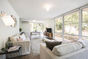 Uma sala de estar moderna com um sofá, uma mesa de centro e uma TV. Uma janela grande oferece vista para o exterior.