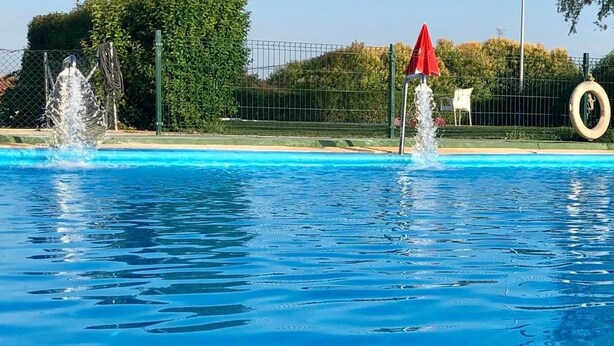 A swimming pool with multiple water jets, a red umbrella, and a tire in the background.