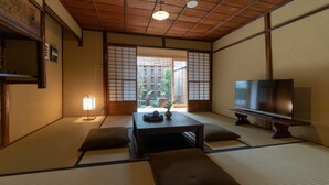 Une pièce de style japonais traditionnel, avec un sol recouvert de tatamis, une table basse, des coussins, un téléviseur et une porte coulissante donnant sur un espace extérieur.