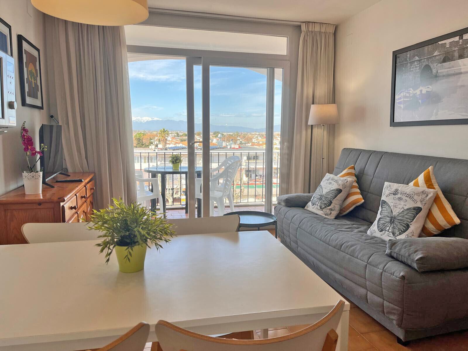 A modern living room with a grey sofa, a dining table, and a balcony with a view of the city.