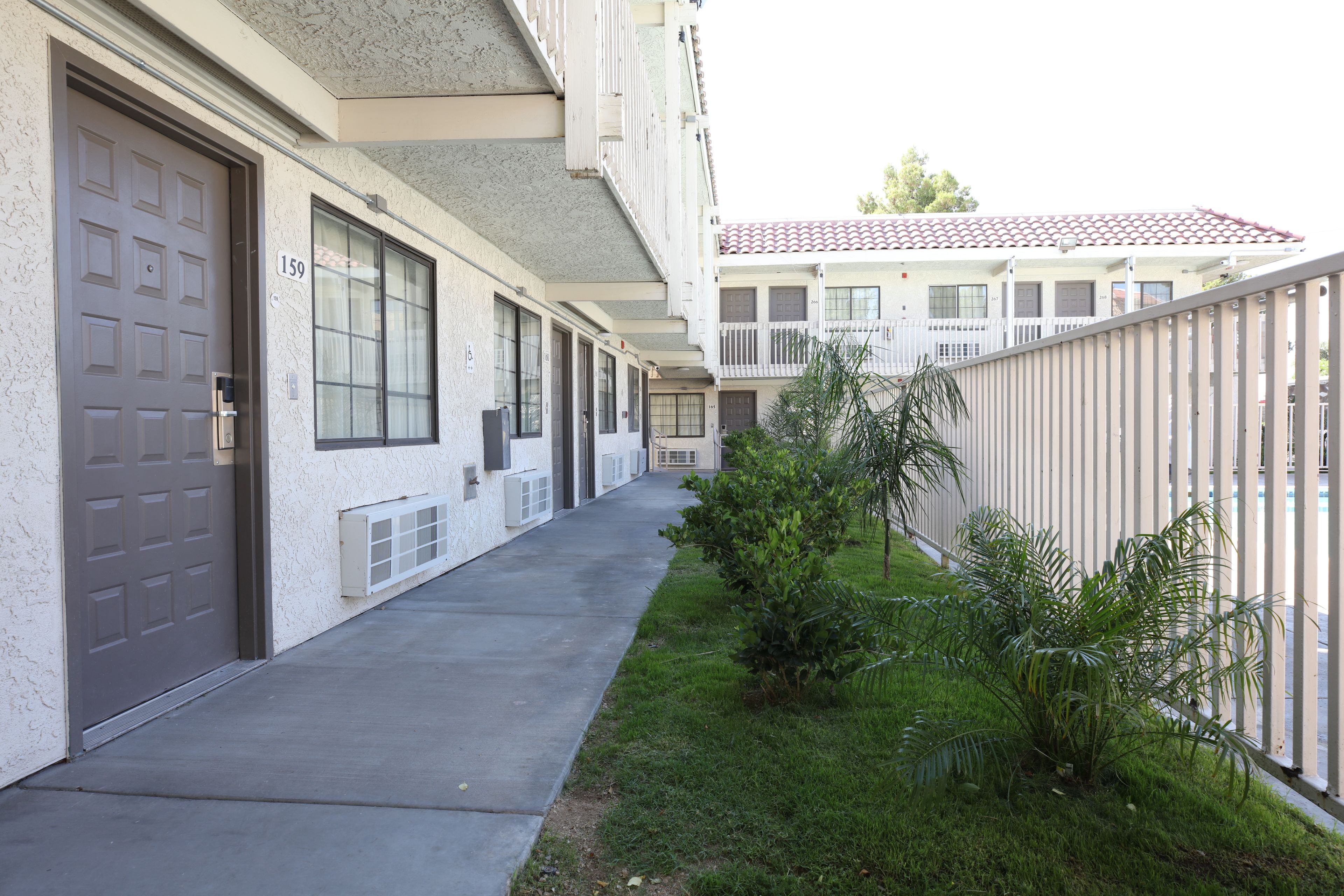 Una hilera de edificios de estilo residencial con números en las puertas, un sendero de concreto y una pequeña zona verde con plantas.