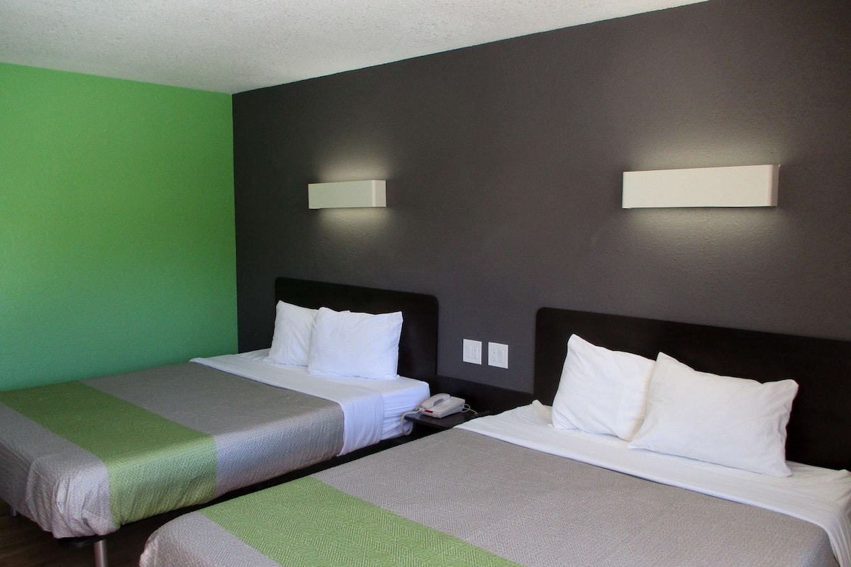 Habitación de hotel con dos camas individuales, cada una con ropa blanca y una colcha verde y gris. Hay lámparas de pared sobre cada cama.