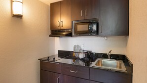 A compact kitchen with dark wooden cabinets, a built-in microwave, a stainless steel sink, and a dish drying rack. - Studio 6 Barstow, CA (Barstow)