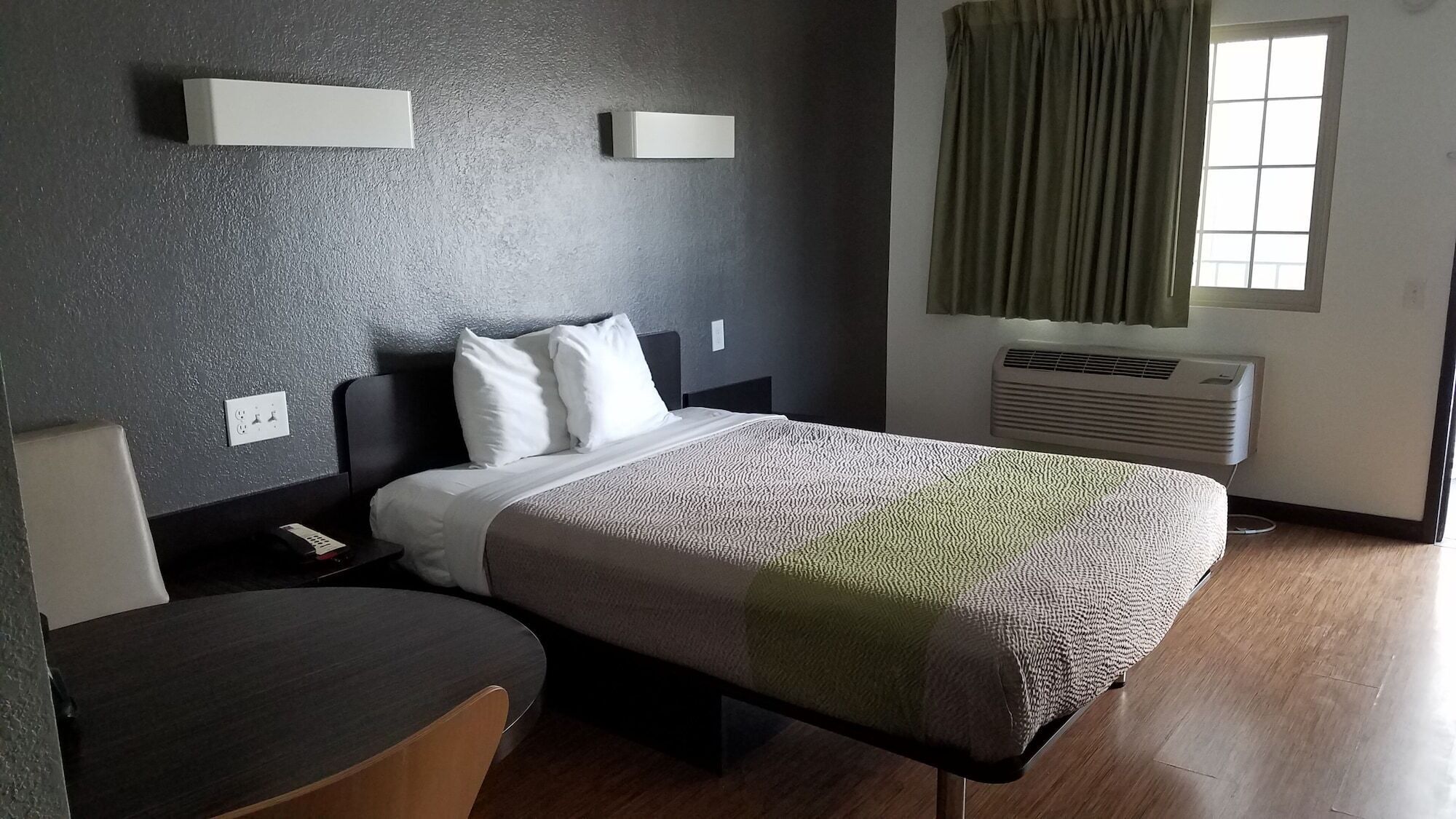 A hotel room with a bed, bedside table, chair, and a window with curtains.