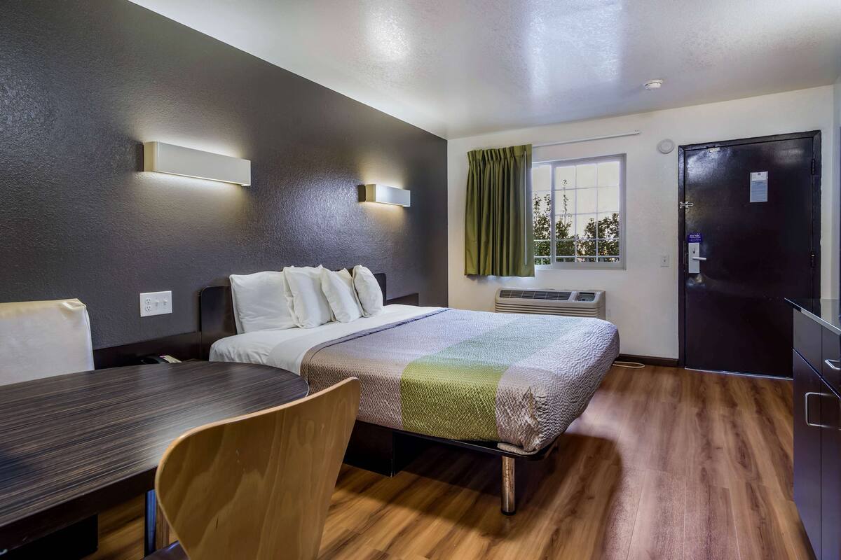 Une chambre d’hôtel comprenant un lit, un bureau, une chaise, une fenêtre avec des rideaux, un appareil de climatisation et une porte.