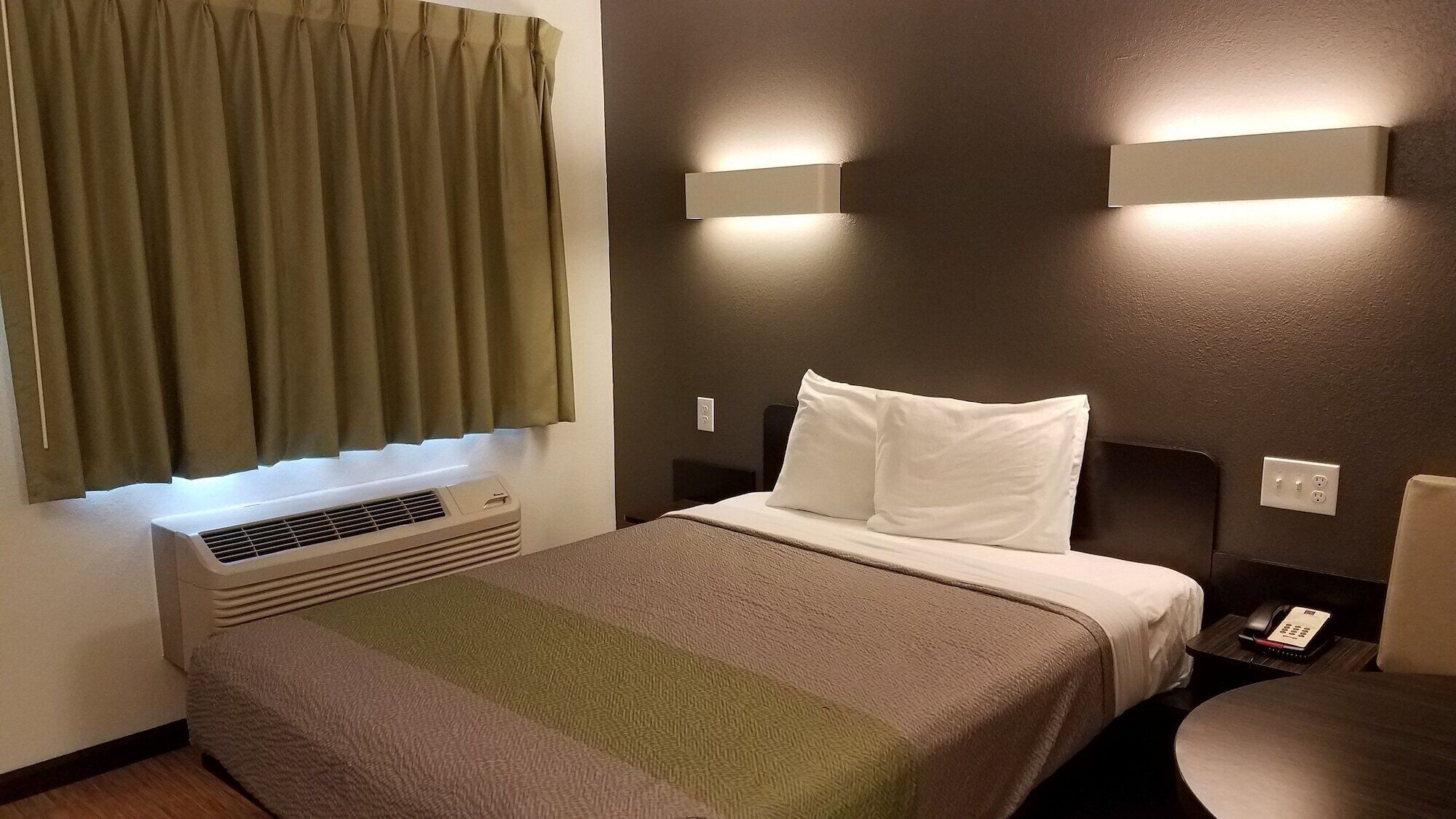 Habitación de hotel con cama, mesita de noche, teléfono, lámpara de pared y ventana con cortinas.