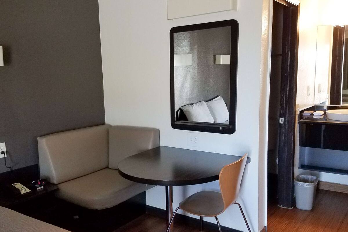 Une chambre d’hôtel compacte comprenant une petite table ronde, une chaise, une banquette intégrée, un grand miroir et une petite poubelle.