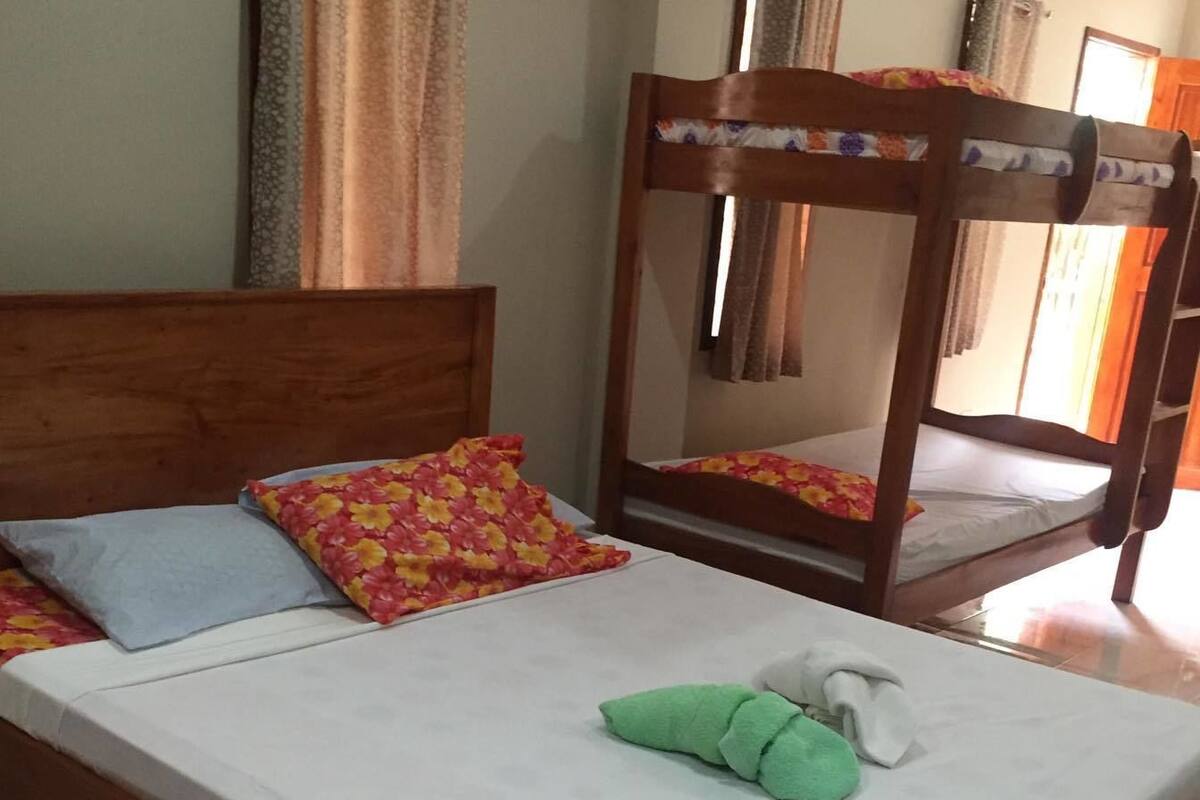 Un lit simple avec un cadre en bois et une literie blanche, agrémenté d’un coussin bleu et d’un coussin à motifs floraux.