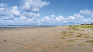 Ein Strand mit weitem Sandbereich, einer kleinen Grünfläche und Blick auf das Meer unter bewölktem Himmel.