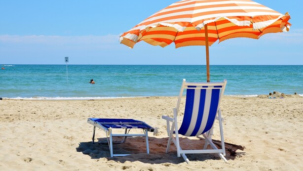 Escena de platja amb un parasol rayat, una cadira de platja blava i blanca rayada, i una cadira de platja blava més petita.