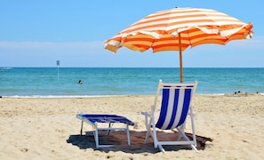 Escena de playa con una sombrilla rayada, una silla de playa azul y blanca a rayas y una silla de playa azul pequeña.