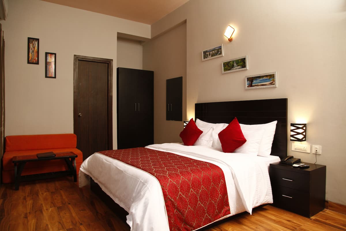 Quarto de hotel com cama, mesa de cabeceira, sofá vermelho e piso de madeira.