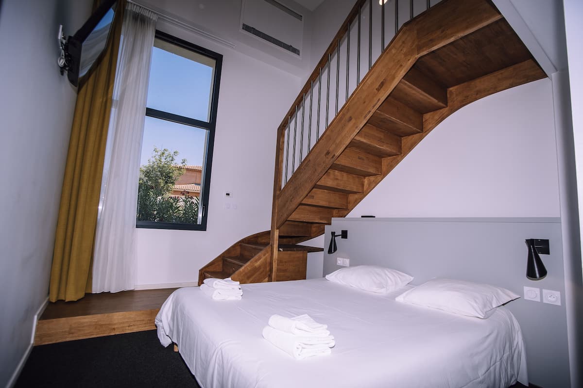 Un dormitorio con una escalera de madera, una cama con ropa blanca y un ventanal con cortinas.