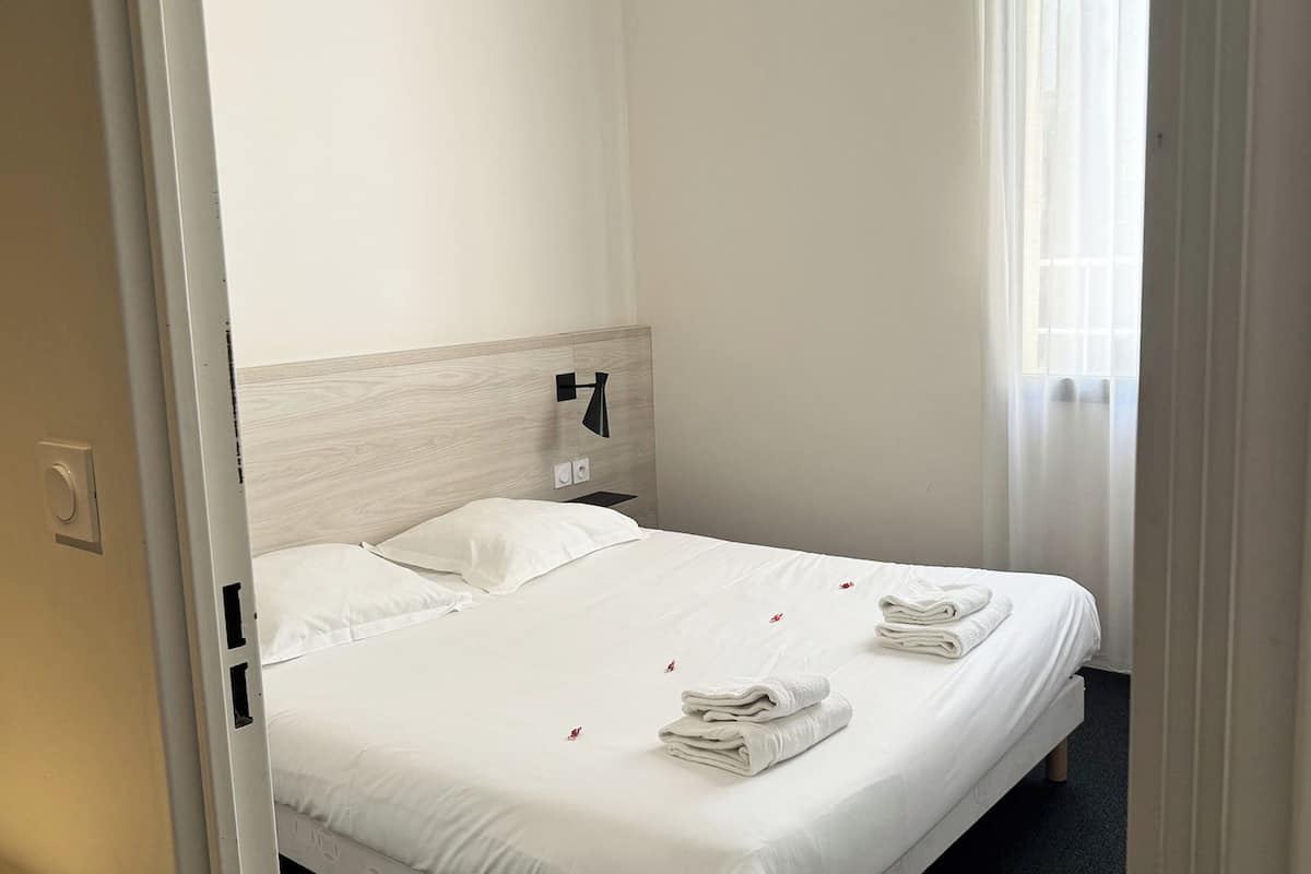 Una habitación de hotel con cama, mesita de noche, lámpara y ventana con cortinas.