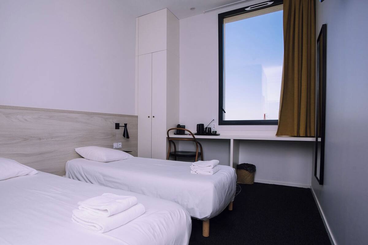 Habitación de hotel con dos camas, un escritorio, una silla, un ventanal con cortinas y un armario.