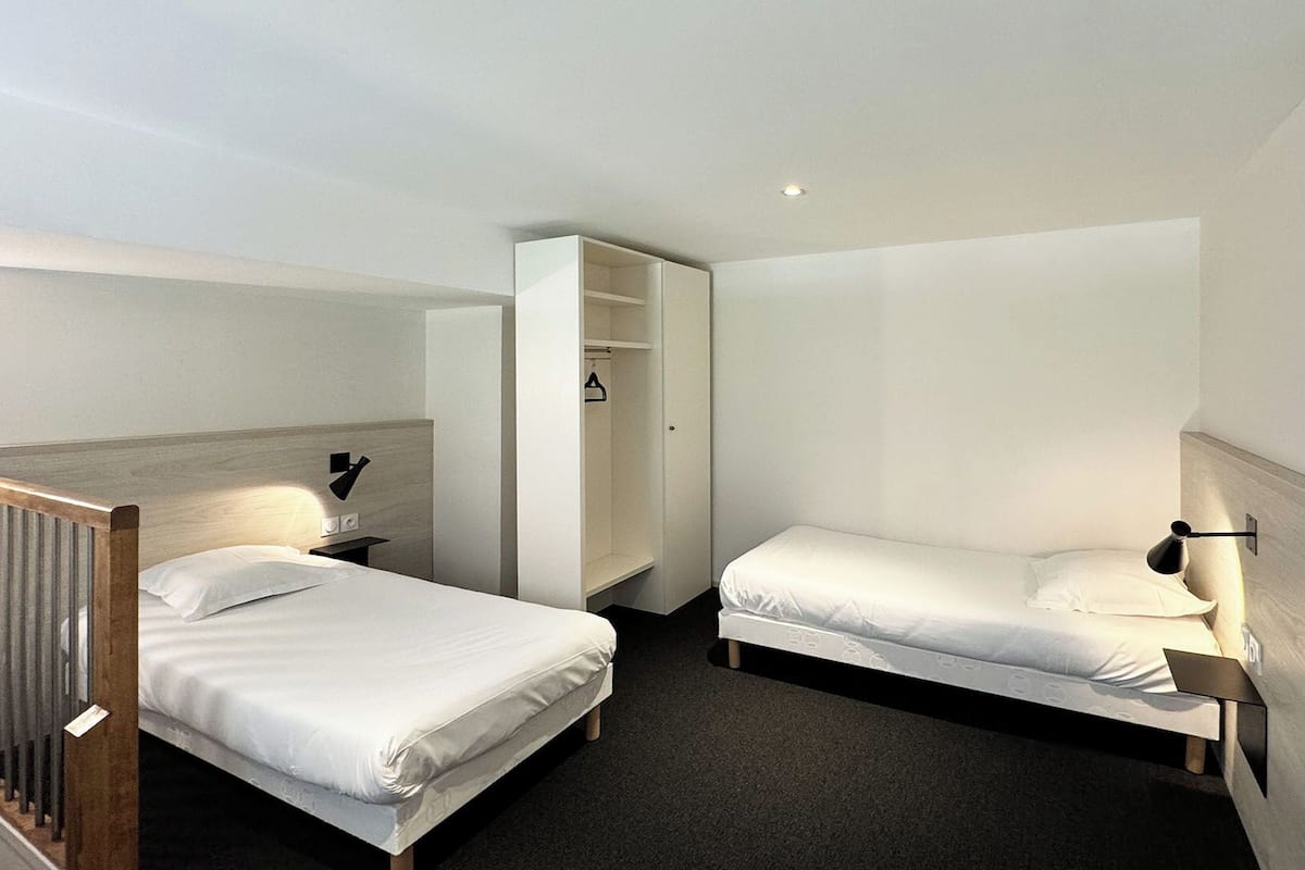 Habitación de hotel con dos camas individuales, un armario, un espejo y un pequeño escritorio con lámpara.