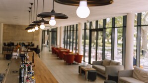 Uma área de bar com várias luminárias pendentes, um balcão comprido com garrafas e assentos em frente a grandes janelas.
