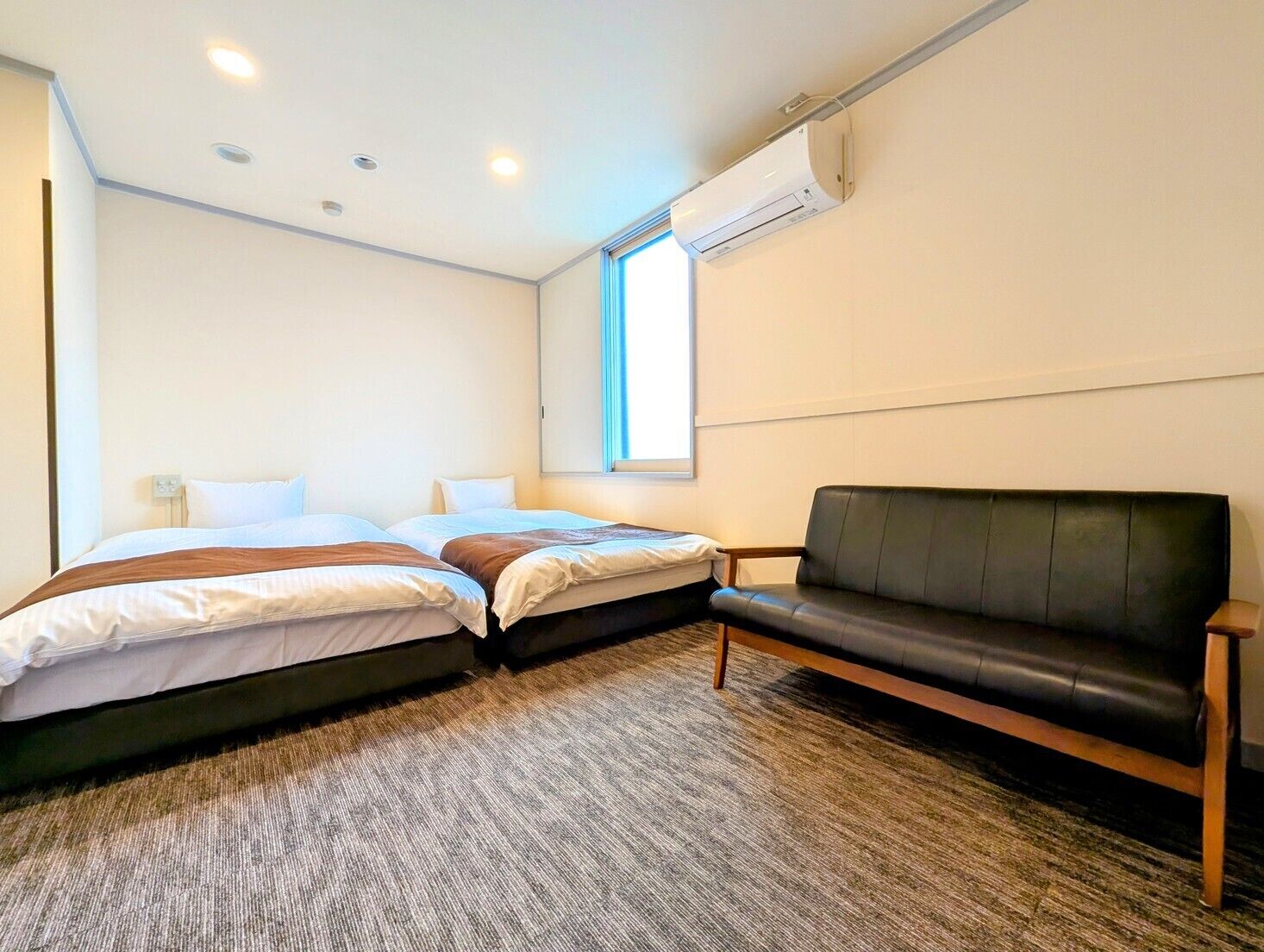Habitación de hotel con dos camas, un sofá de cuero negro, piso alfombrado y un equipo de aire acondicionado en la pared.
