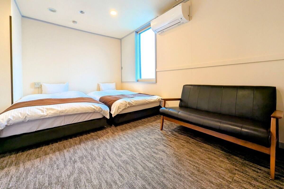 Habitación de hotel con dos camas, un sofá de cuero negro, piso alfombrado y un equipo de aire acondicionado en la pared.