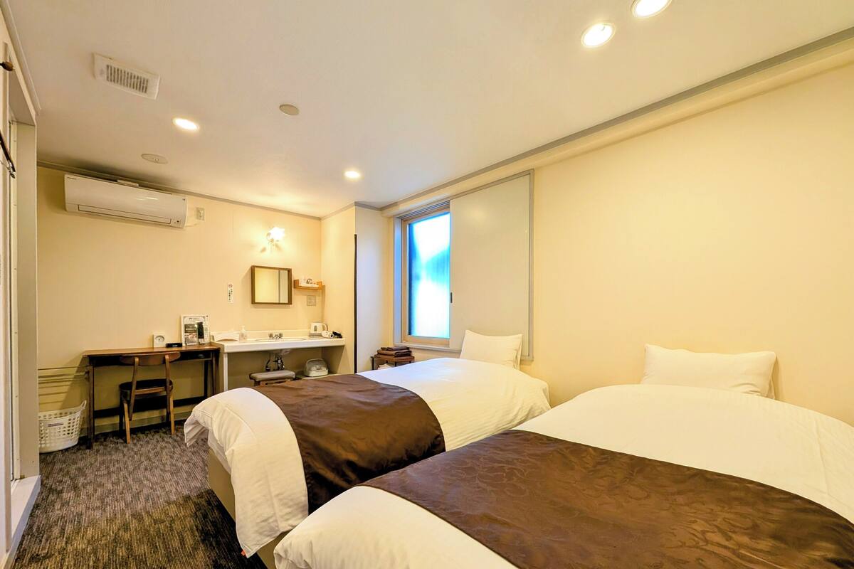 Habitación de hotel con dos camas, un escritorio, una silla, un ventanal y un equipo de aire acondicionado.