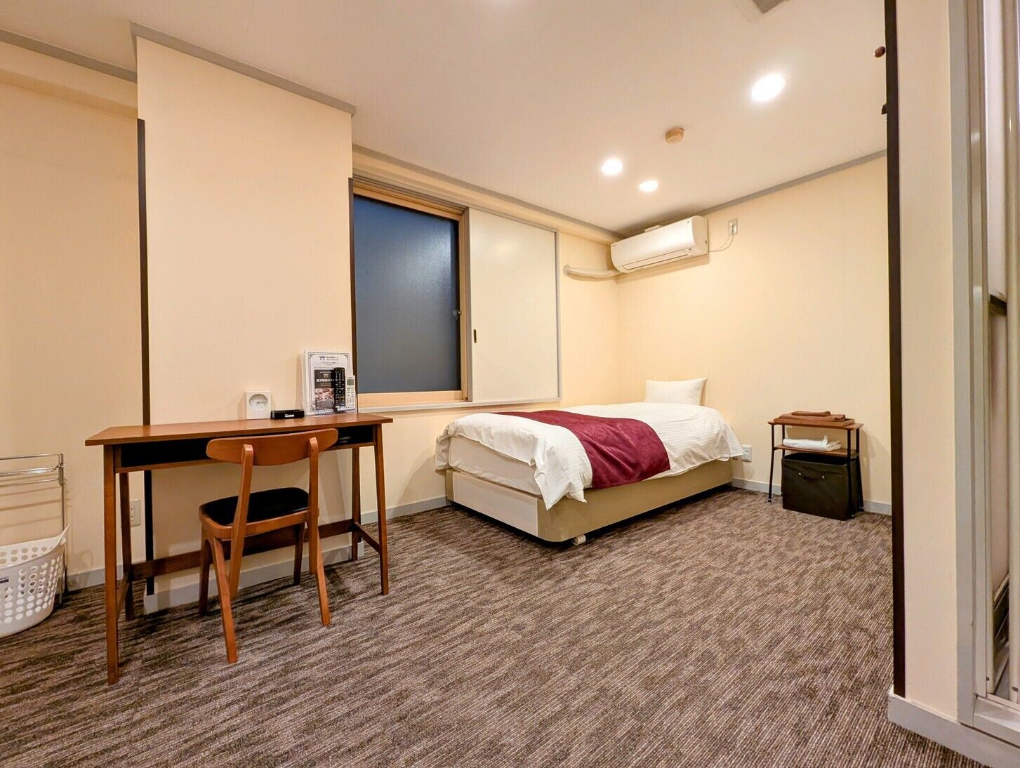 Un dormitorio con cama, escritorio, silla, aire acondicionado y un cubo de basura pequeño.