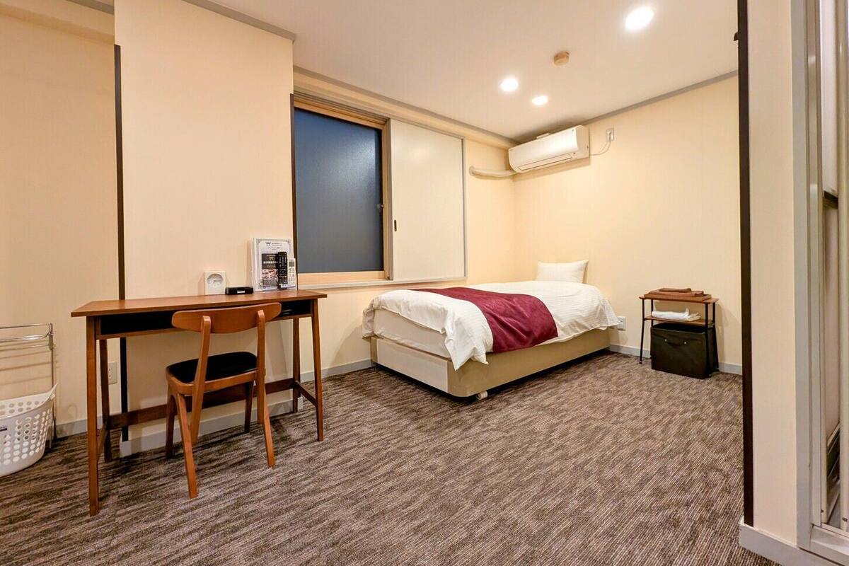 Un dormitorio con cama, escritorio, silla, aire acondicionado y un cubo de basura pequeño.