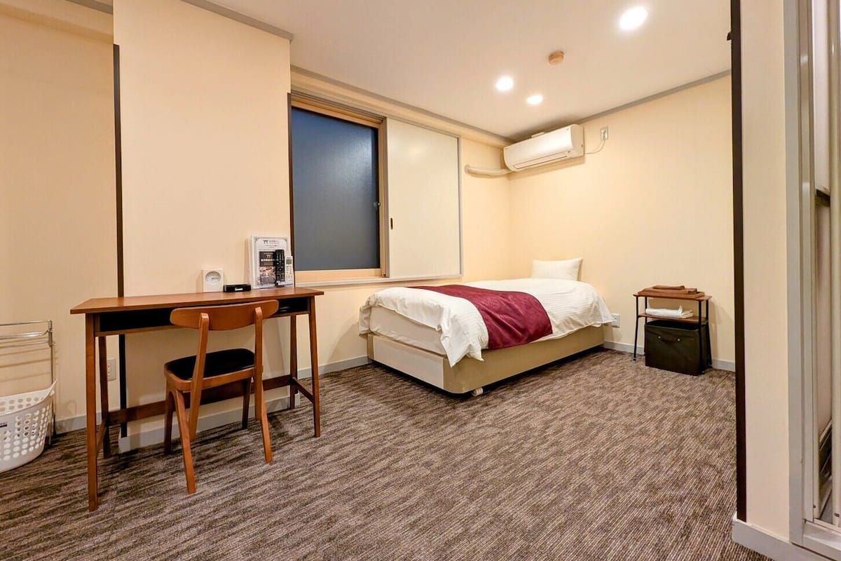 A bedroom with a bed, a desk, a chair, an air conditioner, and a small trash bin.