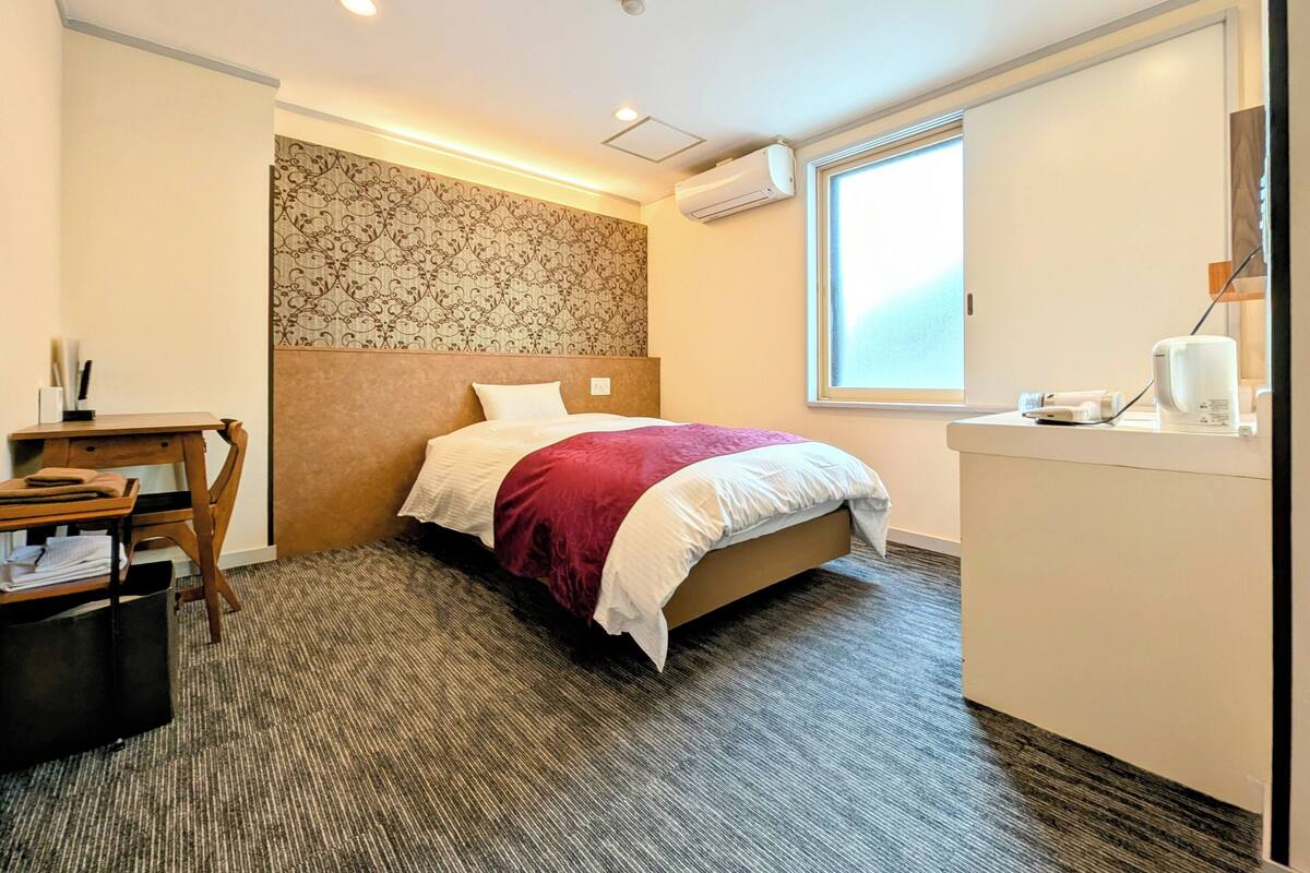 Habitación de hotel con cama, escritorio, silla, mesita, espejo y ventana.