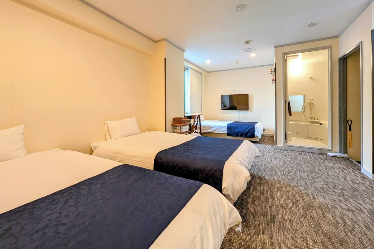 Habitación de hotel con dos camas, televisor, baño con espejo y puerta que da a otra habitación.