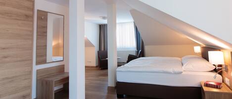 A modern hotel room with a sloped ceiling, a large bed with white linens, a bedside table with a lamp, a wooden bench, and a large mirror on the wall.