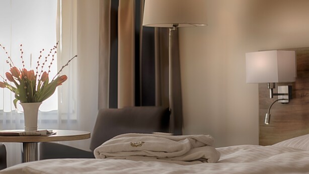 Habitación de hotel con cama, una silla, una mesa con un florero y una lámpara, además de una lámpara de pared.