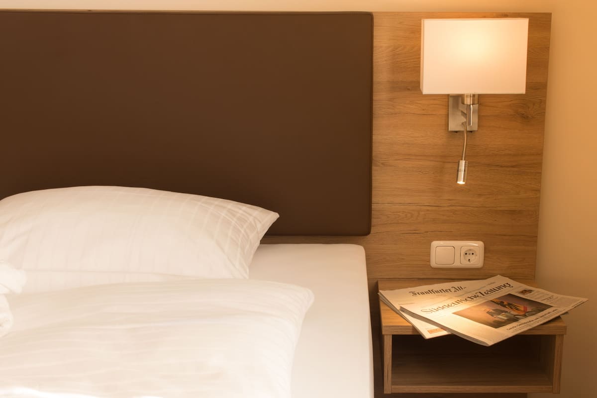 A neatly made bed with white linens, a wooden headboard, a bedside lamp, and a nightstand with a newspaper.