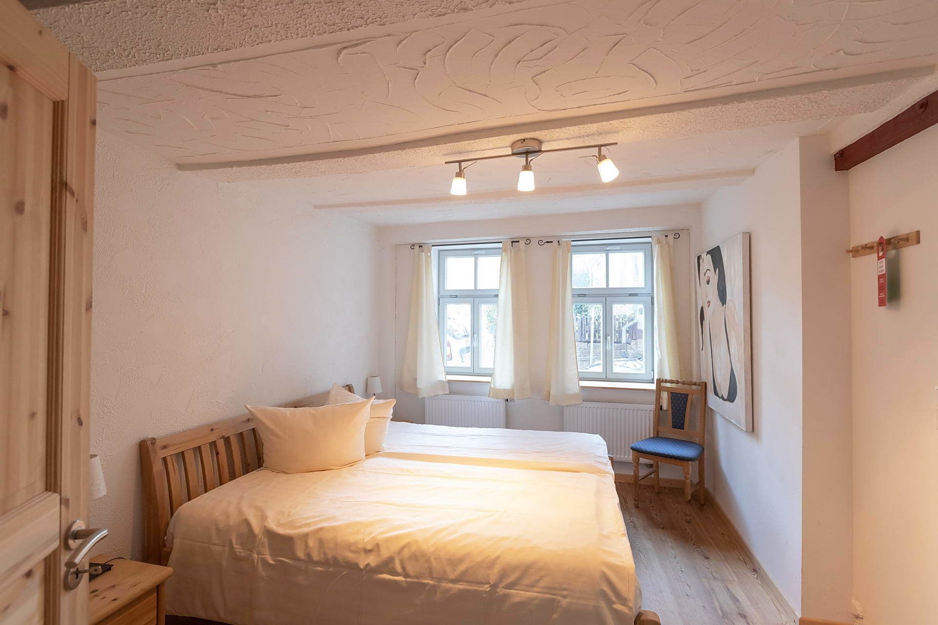 Un dormitorio con cama, una silla, una ventana con cortinas y un cuadro en la pared.