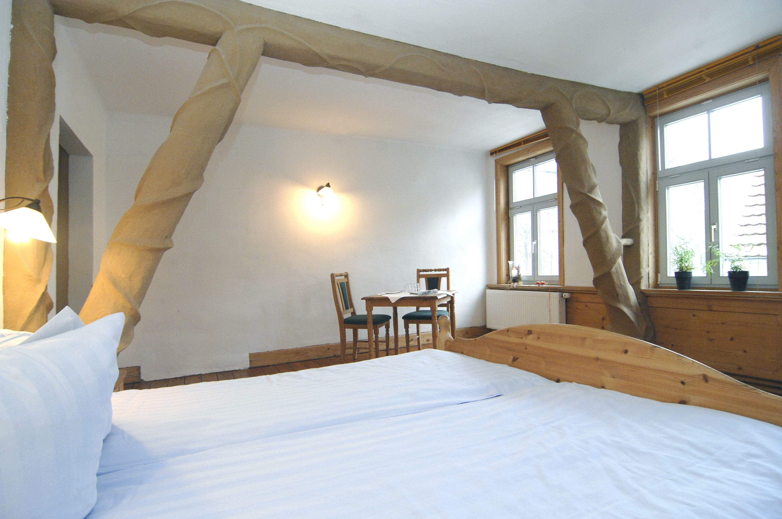A bedroom with a wooden bed, a desk with chairs, a window with plants, and a wall-mounted light.