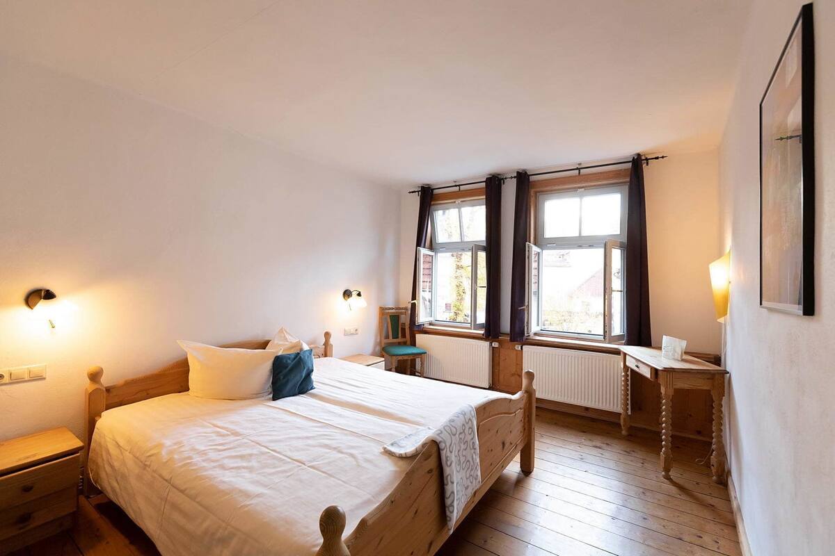 Une chambre à coucher avec un lit en bois, deux fenêtres avec des rideaux, une table de chevet et un radiateur.