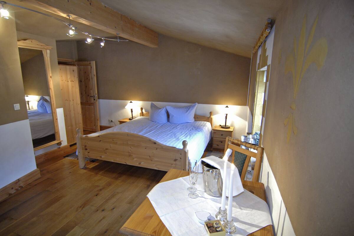 Un lit en bois dans une pièce dont le sol est également en bois, une table de chevet avec une lampe et un miroir accroché au mur.