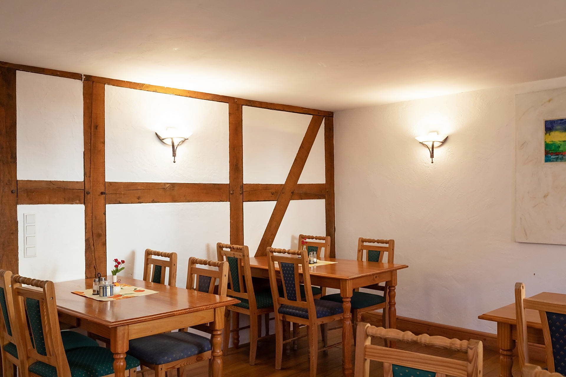 Un espace repas avec des tables et des chaises en bois, deux lampes fixées au mur et un mur blanc agrémenté d’une structure en poutres de bois.