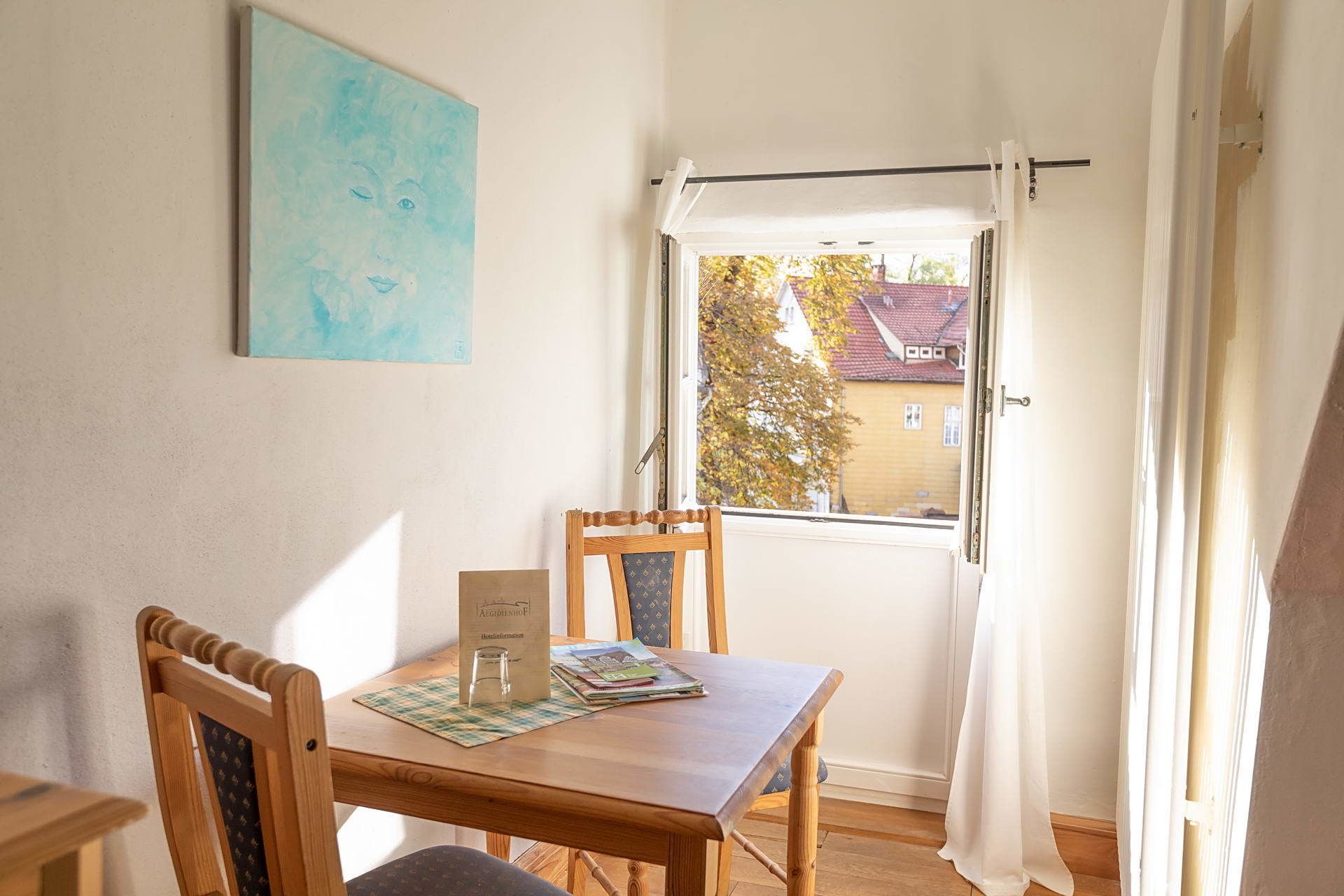 Una habitación con una mesa de madera, dos sillas, un ventanal con vistas a un edificio y árboles, y un cuadro en la pared.