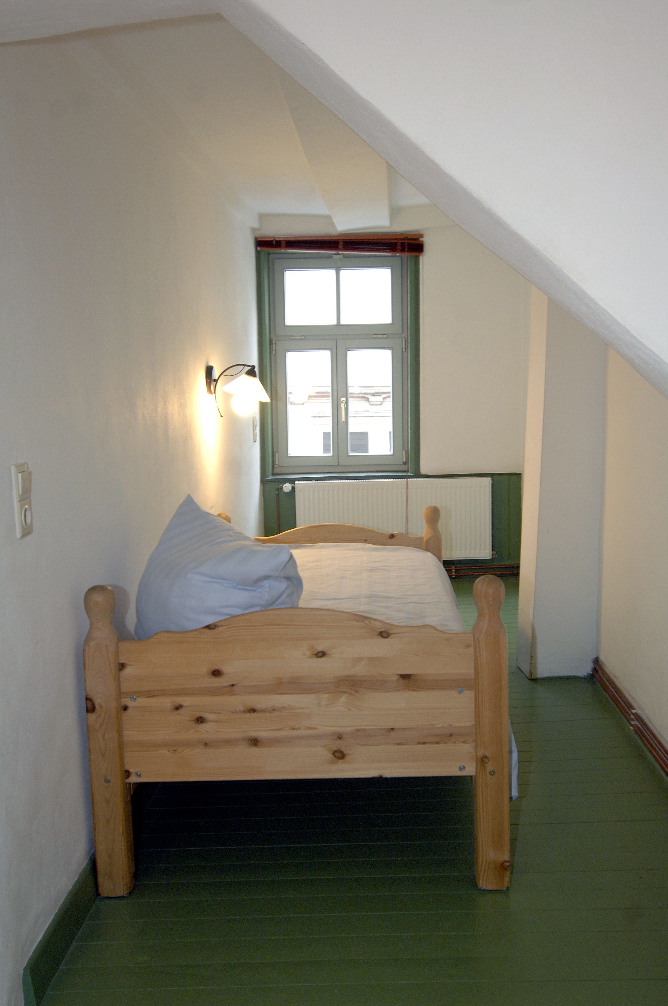 A small, single-bed room with a sloped ceiling, a window with a view, and a wall-mounted light fixture.