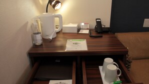 Ein Hotelzimmer mit Schreibtisch aus Holz, einer Kaffeemaschine, einer Lampe, einem Telefon und einer Schuhputzgelegenheit.