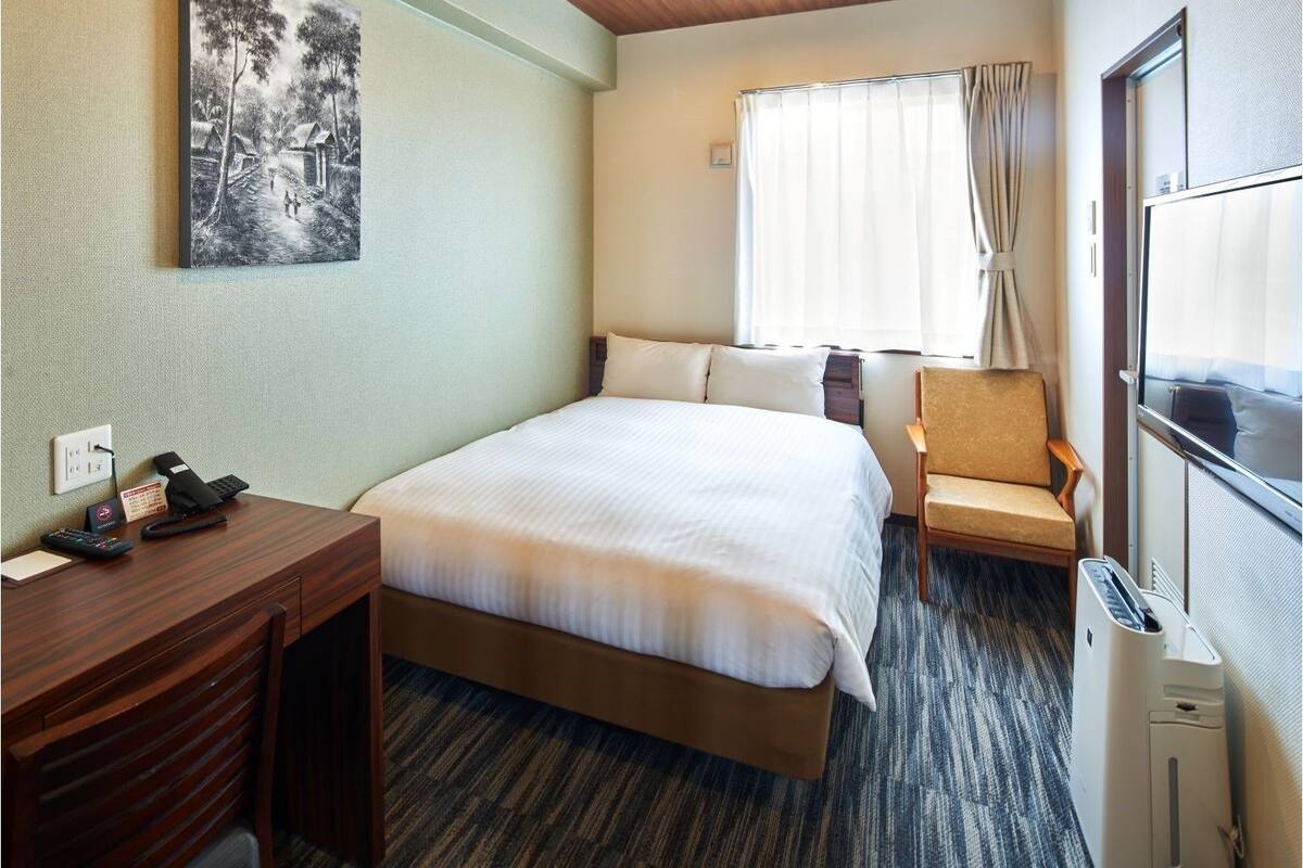 Ein Hotelzimmer mit Bett, Schreibtisch mit Telefon, Stuhl, Fenster mit Vorhängen und Heizkörper.