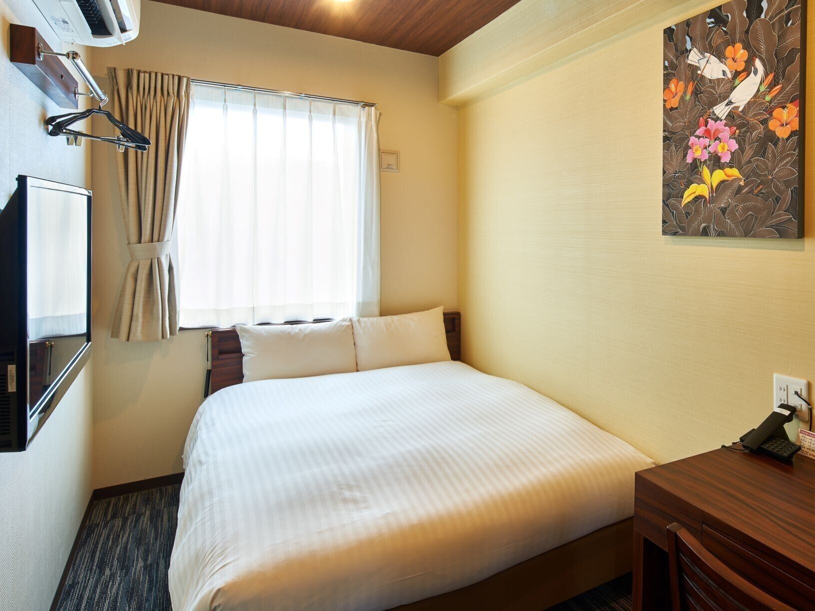Quarto de hotel com cama, escrivaninha, televisão, aparelho de ar condicionado, janela com cortinas e quadro pendurado na parede.