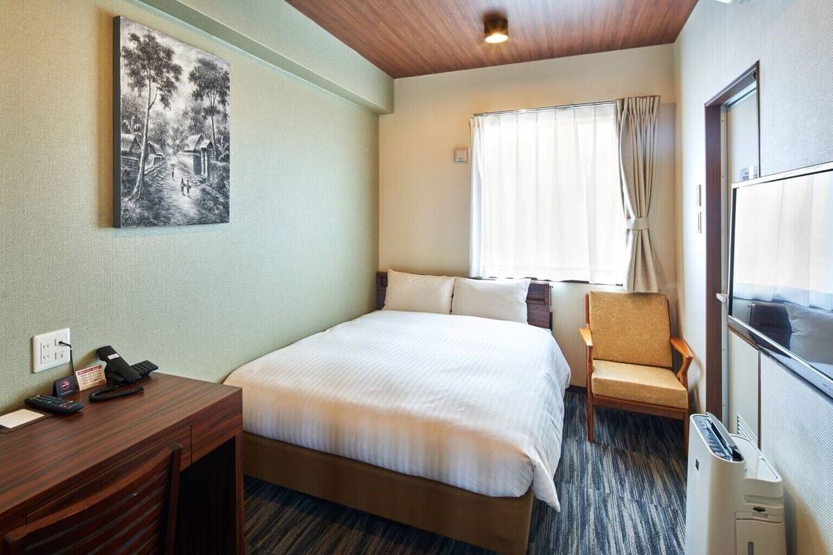 Ein Hotelzimmer mit Bett, Schreibtisch mit Telefon, Stuhl, Fenster mit Vorhängen und Heizkörper.