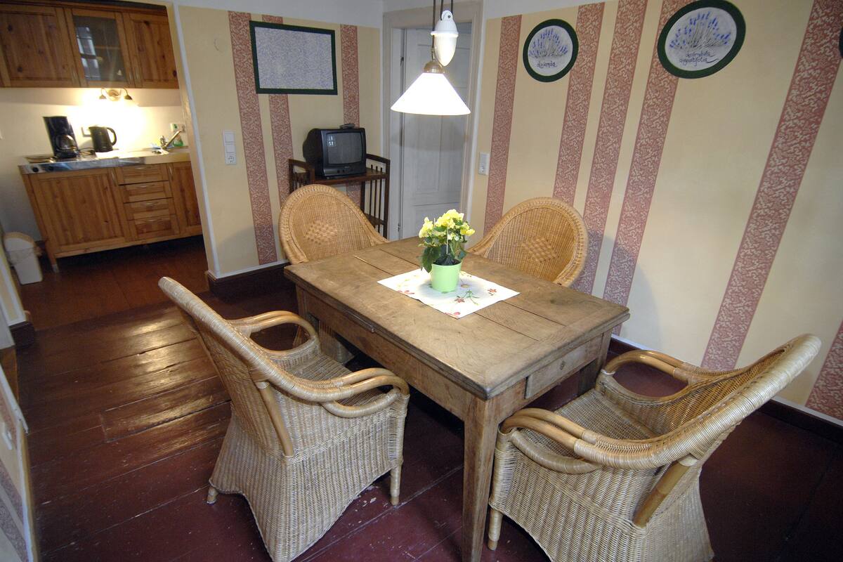 A dining area with a wooden table, wicker chairs, a small green vase with flowers, and a television in the background.