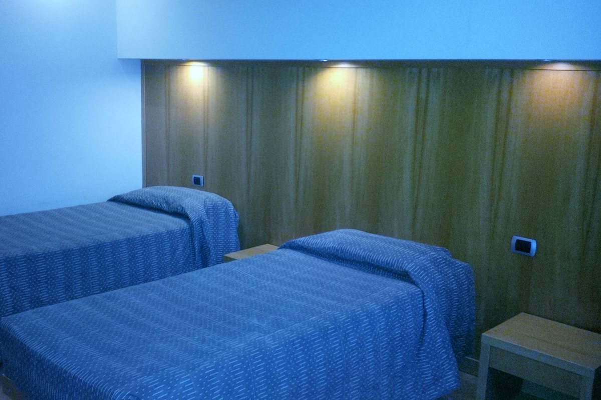 Una habitación con dos camas individuales, una mesita pequeña y una lámpara fijada a la pared.