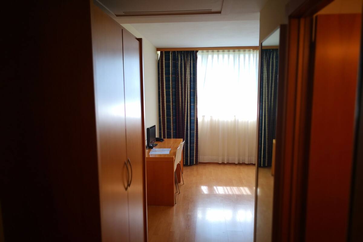 Una habitación con escritorio de madera, una silla, televisor y ventana con cortinas.