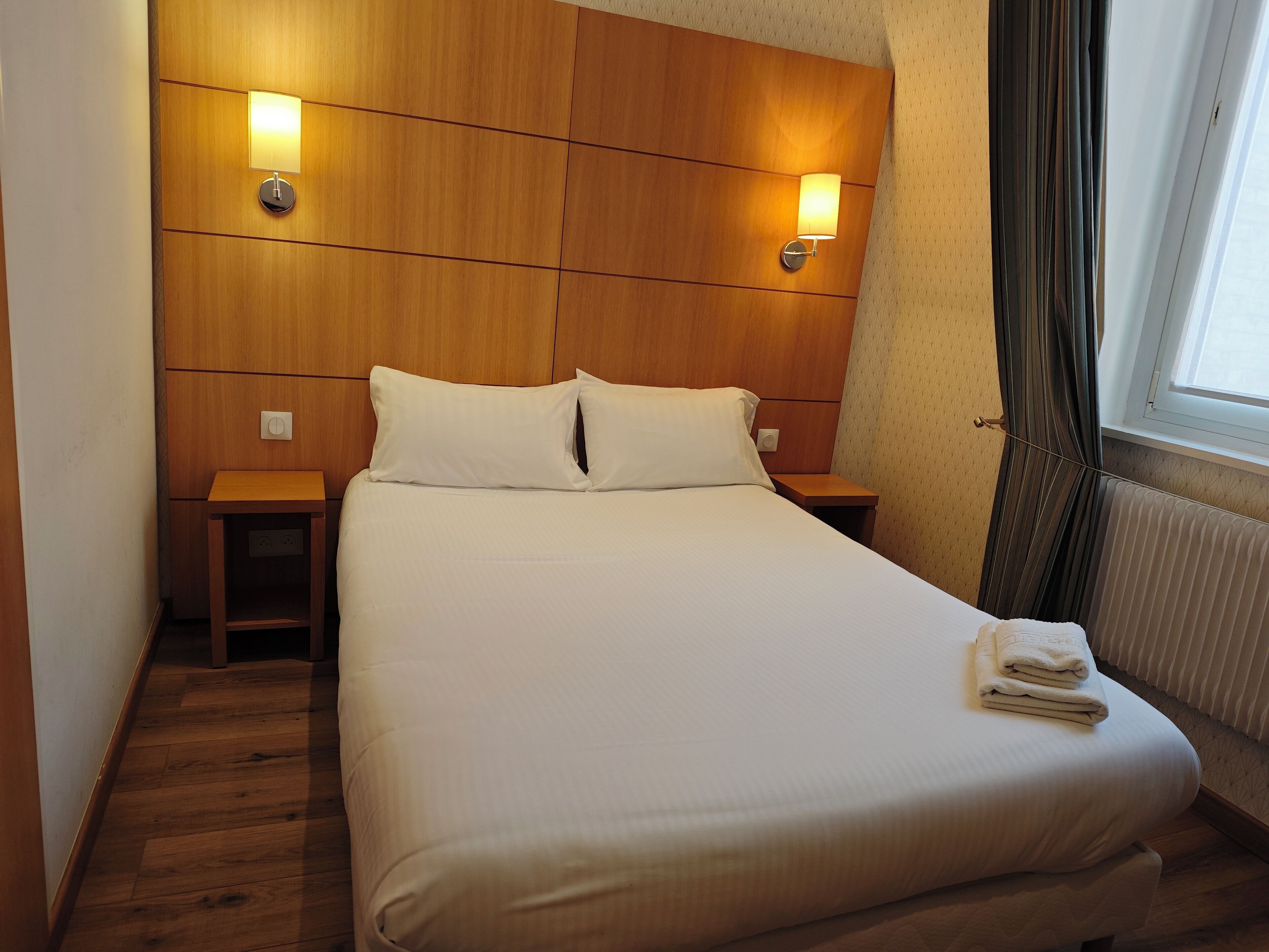 A neatly made bed with two pillows, wooden headboard, bedside lamps, and a window with curtains.