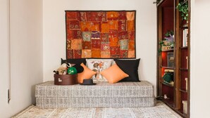 A cozy seating area with a patterned tapestry, a small table with a hat and a green plant, and a bookshelf with books and plants. - Nephiria's Art House, Under the Acropolis (Athens)