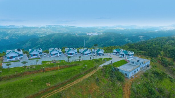 A cluster of modern, blue-roofed houses with a central building, surrounded by lush greenery and mountains in the background. - Kananam Retreat (Thodupuzha)