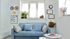 A blue sofa with patterned pillows, a small white table with a book, and a window with decorative baskets and plants. - Nephiria, Under the Acropolis, In the heart of Plaka (Athens)